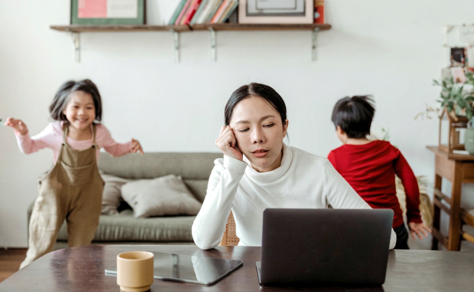 A woman working on a laptop at home while her two kids run around the living room